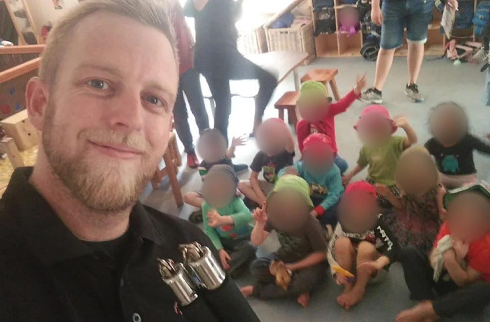 A man with a beard taking a selfie with a group of children sitting on the floor behind him, some wearing colorful hats, inside a room with shelves and benches. Children’s faces blurred for privacy.