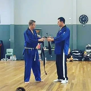 Two martial artists in blue uniforms, one with a white belt and the other with a black belt, exchanging a trophy in a gym or dojo.