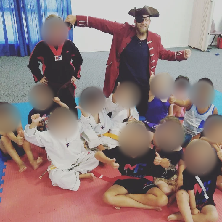 Children in martial arts uniforms and casual clothing posing with two adults in costume, inside a room with blue curtains and gray carpet, some children giving thumbs-up and smiling. Children’s faces blurred for privacy.