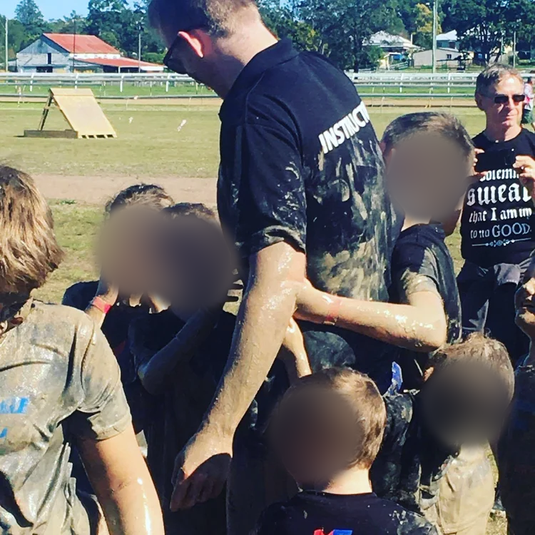 Group of children and adults outdoors, covered in mud, celebrating after a muddy event or activity, with a grassy field in the background. Children’s faces blurred for privacy.