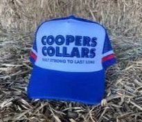 A blue and white Coopers Collars baseball cap with purple stripes on the side, placed on gravel ground.