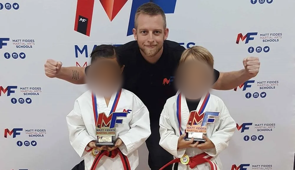 Dream Kids Australia Founder Adrian Anderson MMA Instructor Two young children in martial arts uniforms holding trophies and medals, standing with an adult man in a martial arts school with a branded backdrop. Children’s faces blurred for privacy.