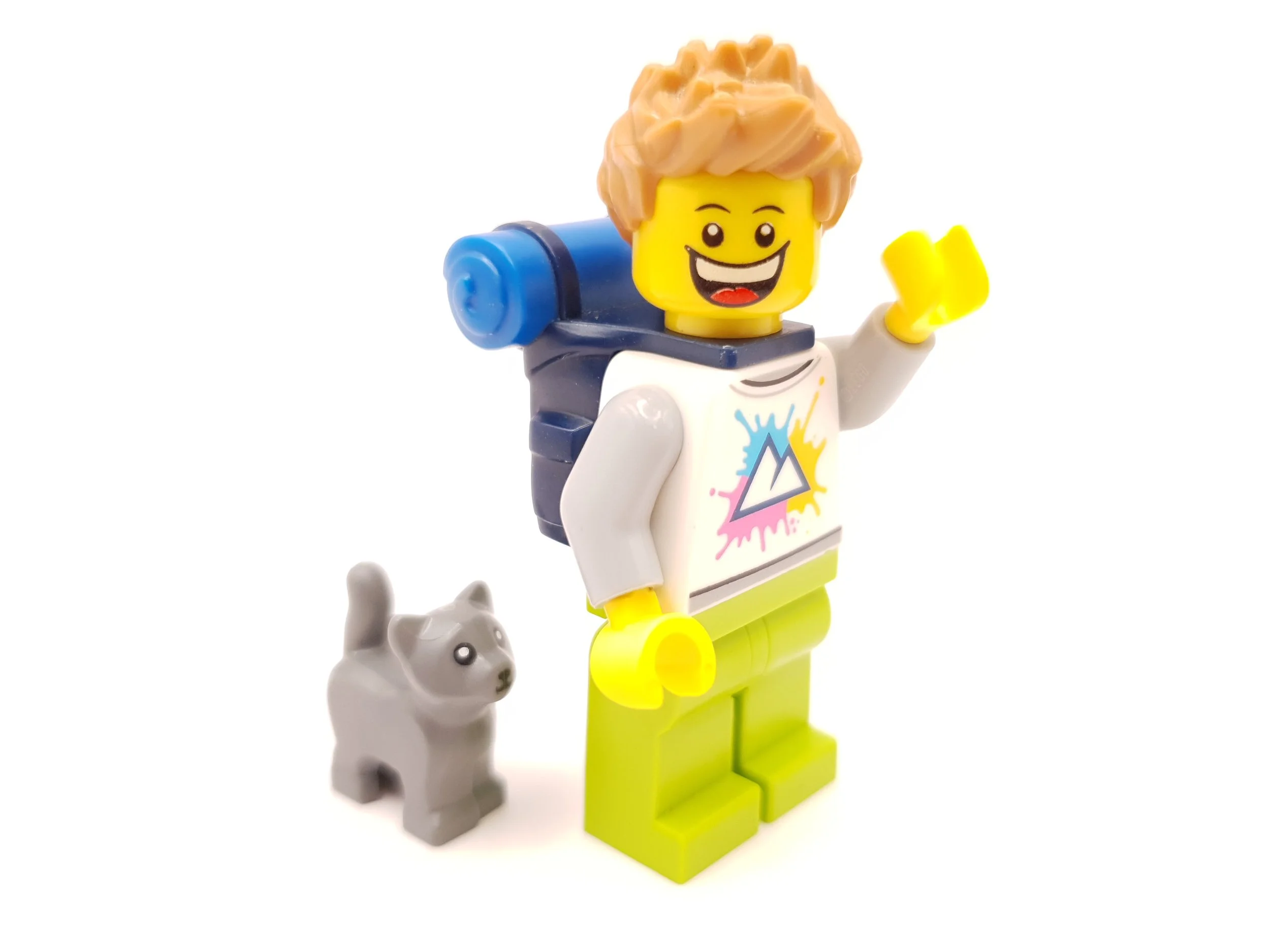 Lego minifigure of a smiling person with yellow skin, brown hair, wearing a white sweatshirt with colorful splash and triangle design, yellow legs, blue backpack, and a small gray dog figure, all on a white background.