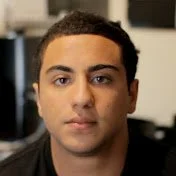 superwog1 youtube logo Close-up of a young man with short curly hair and brown eyes, facing the camera. Background is blurred.