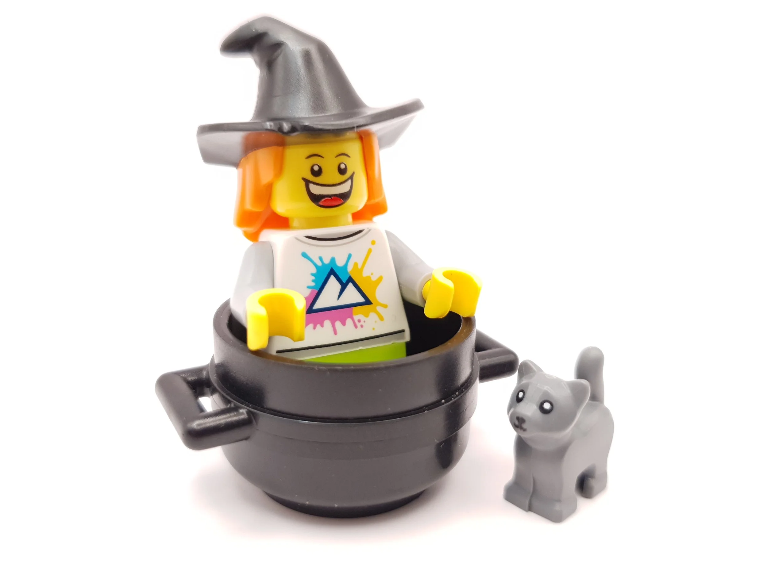 Lego witch figure with orange hair, wearing a gray hat, in a black cauldron, accompanied by a small gray cat figure, against a white background.