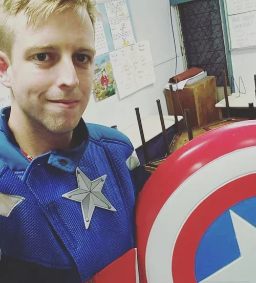 A man with light hair and a slight smile wearing a blue shirt with captain America cosplay costume, standing next to a Captain America shield in a classroom with children's drawings and tables in the background.