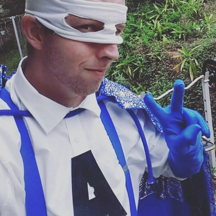 A man dressed in a costume resembling a character from the movie 'G.I. Joe' with a white mask covering his eyes, white shirt with a black letter 'A', and blue accents, making a peace sign gesture outdoors with greenery in the background.
