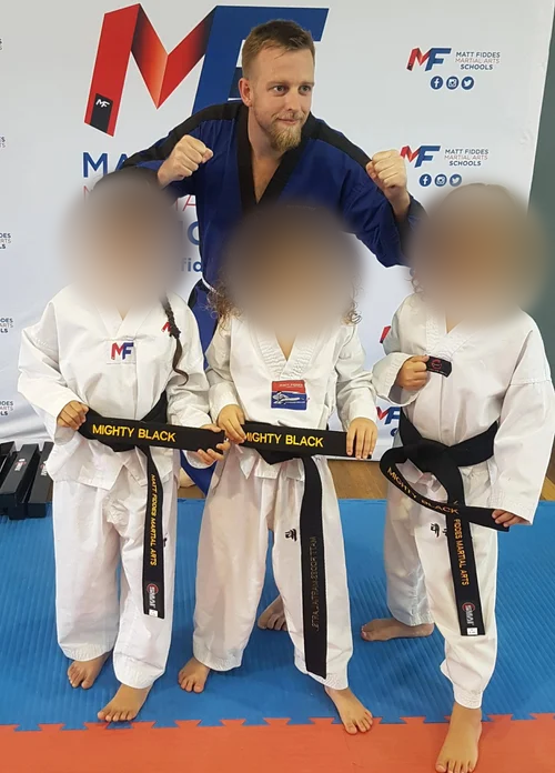 Three young girls in martial arts uniforms with black belts standing on a blue and red mat, holding black belts with yellow text. Behind them, a man in a blue martial arts uniform stands with his fists raised in a fighting stance. The backdrop displa