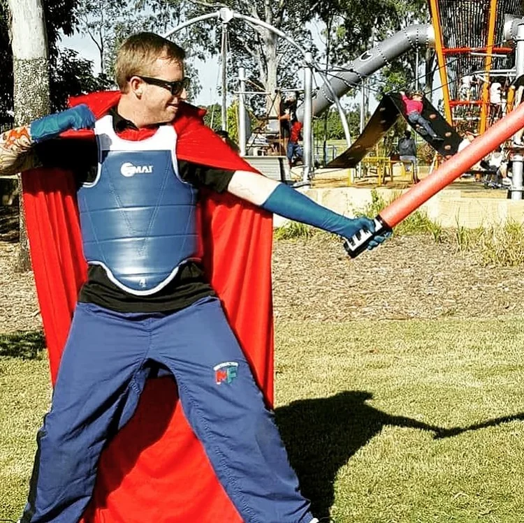 Child dressed as a superhero with a blue chest armor, black shirt, blue pants, red cape, and sunglasses, wielding a foam sword in a playground.