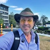 WalkAboutWithRob youtube logo Man wearing a hat, glasses, and a backpack, smiling outdoors in a city environment.