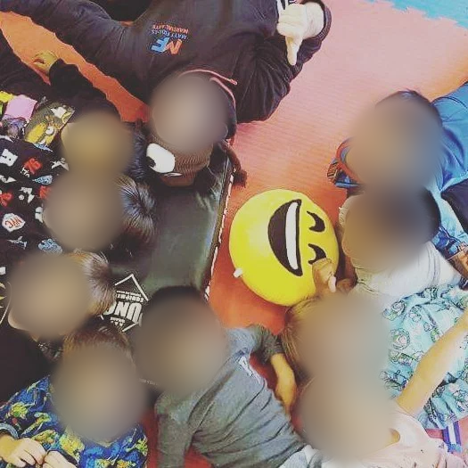 A group of children lying on the floor around a large yellow emoji pillow with a smiling face, all smiling and looking up at the camera. Children’s faces blurred for privacy.
