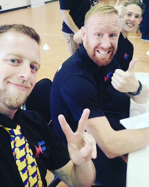 Two men sitting at a table taking a selfie, smiling and making peace and thumbs-up gestures, with two women in the background giving thumbs-up sign.