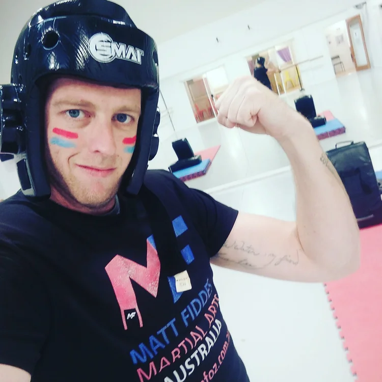 A young man wearing a black helmet and a black T-shirt, flexing his bicep and smiling at the camera, with face paint in red and blue stripes on his cheeks, in a martial arts or gym training facility.