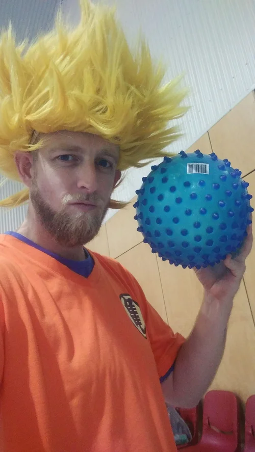 A man dressed in an orange sports jersey with a yellow spiky wig and a goatee, holding a blue spiked ball, standing indoors against a beige wall with a partial view of red chairs.