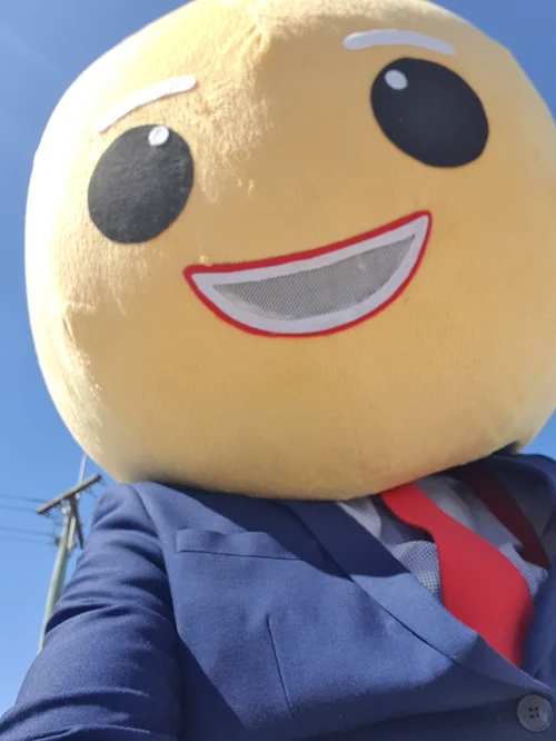 Person wearing a large, round emoji mascot costume with a smiling face, big eyes, white eyebrows, and a red outline mouth, outdoors in a blue sky with power lines in the background.