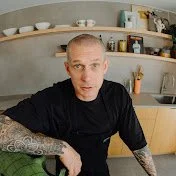Andy Cooks Youtube logo Man with tattoos on his arm wearing a black shirt sitting in a modern kitchen