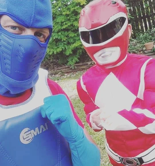 Two individuals dressed in colorful superhero costumes outdoors, with greenery in the background. One wears a blue mask and gloves, and the other wears a red helmet and costume.