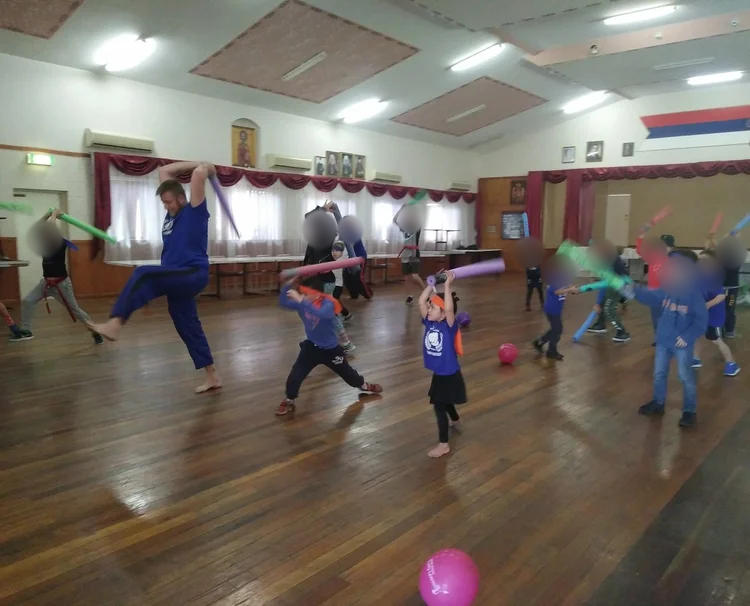Children participating in a martial arts class, using foam batons, in a large wooden-floored room with red curtains and framed pictures on the wall. Children’s faces blurred for privacy.