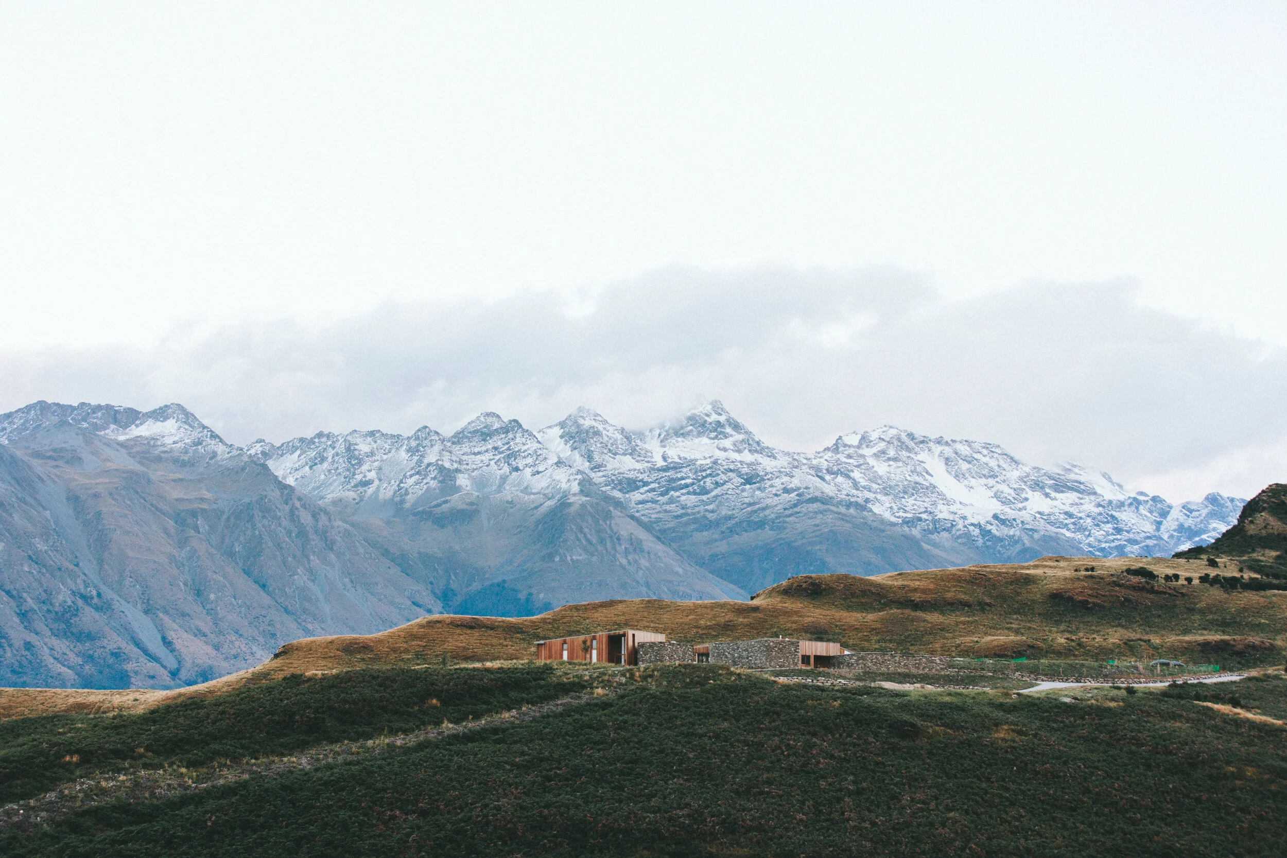 Paysage de montagnes avec sommets enneigés, collines verdoyantes, et une maison moderne en bois au premier plan.