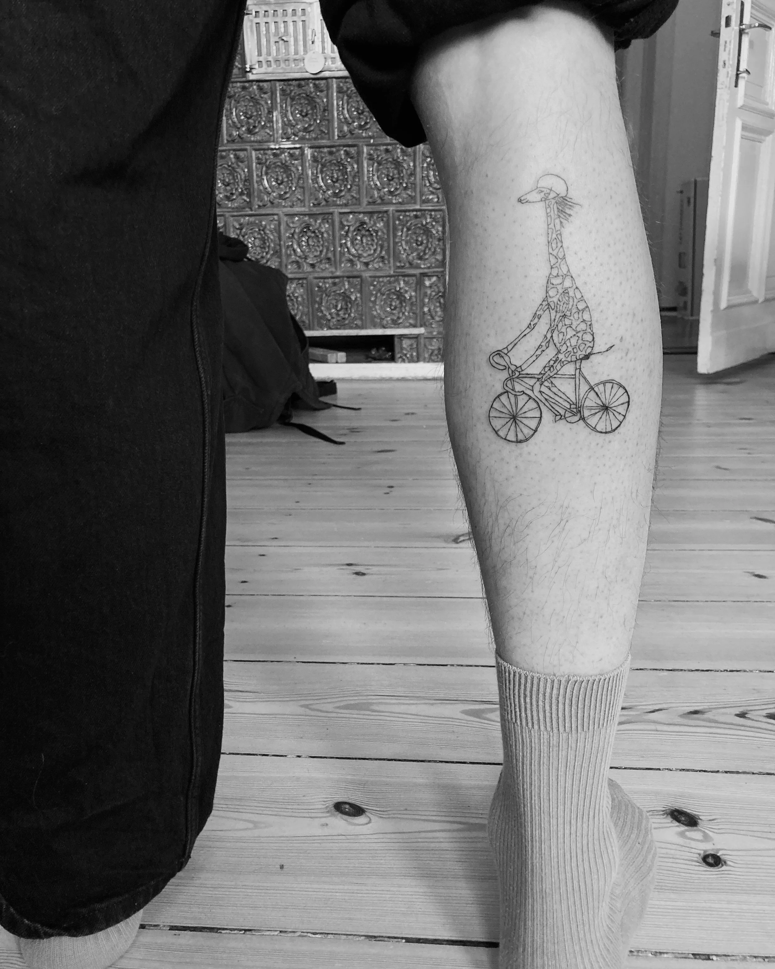 Finelinetattoo von Rahel Bachem auf einem Bein. Motiv: eine fahrradfahrende Giraffe mit Helm