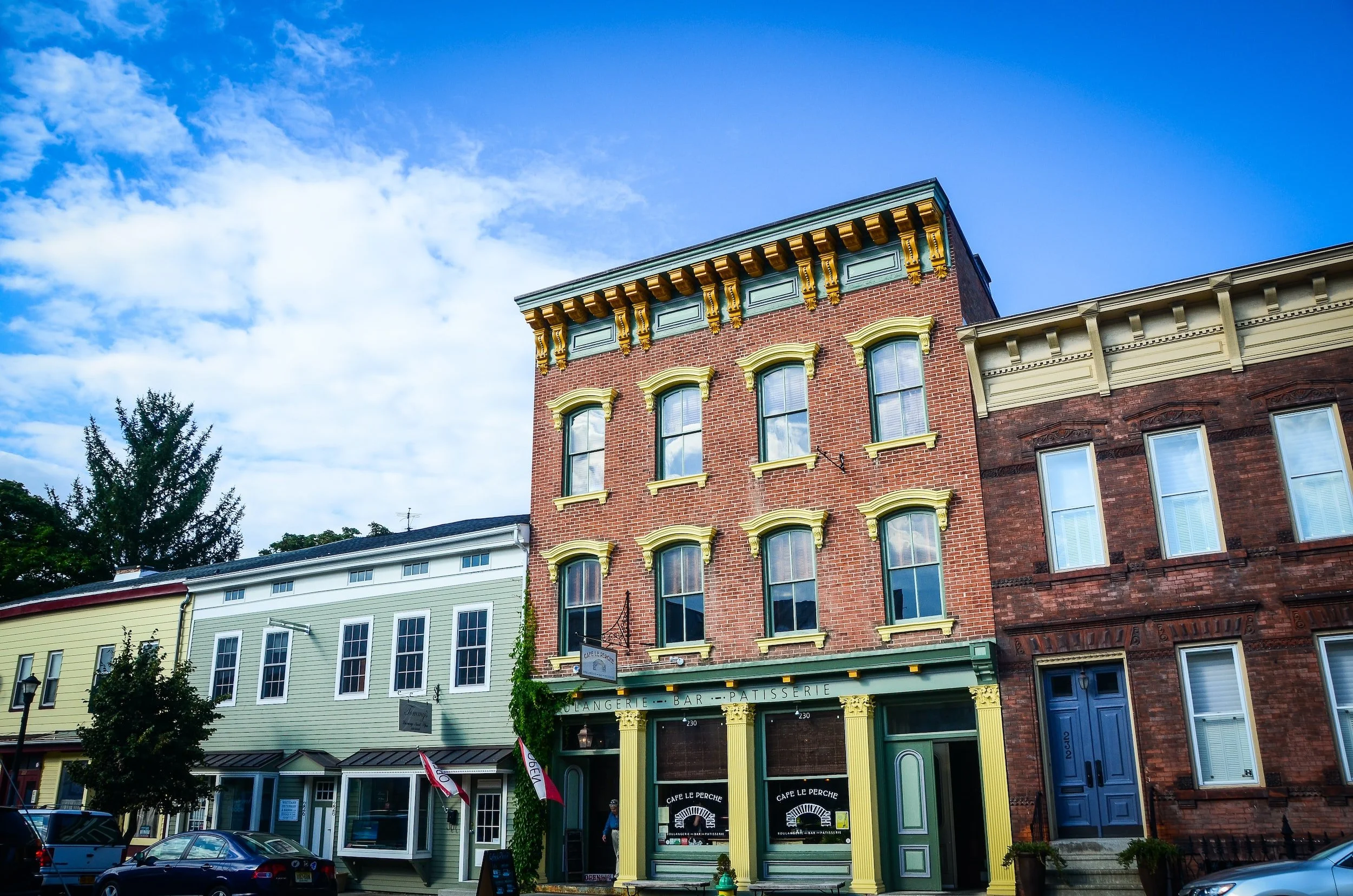 Colorful brick buildings with storefronts on a street under a blue sky with clouds, including a cafe with a green and yellow facade.