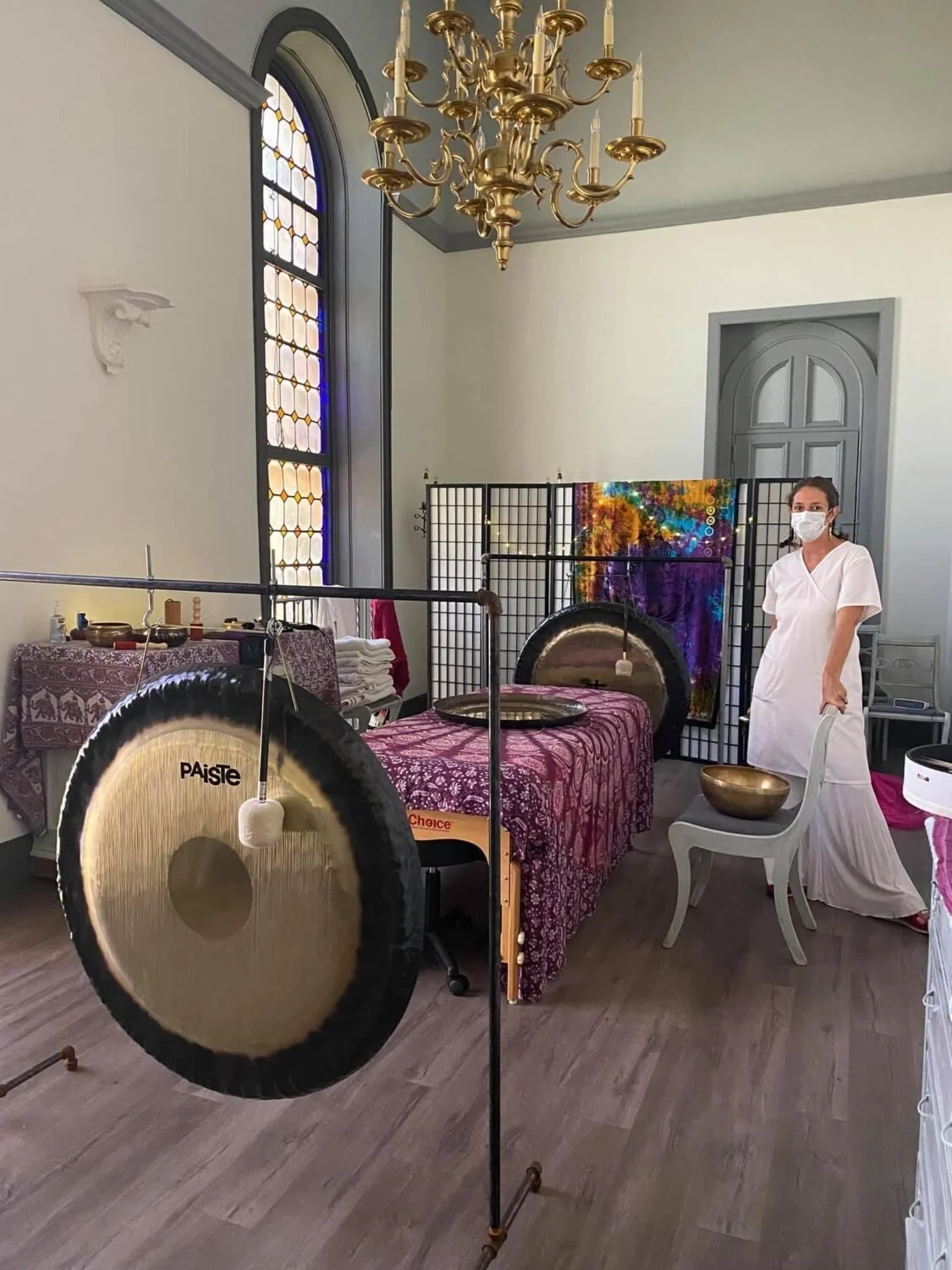 A woman wearing a face mask and white dress standing next to a Tibetan singing bowl in a room with large stained glass window, chandelier, and sound healing setup including gongs and other instruments.