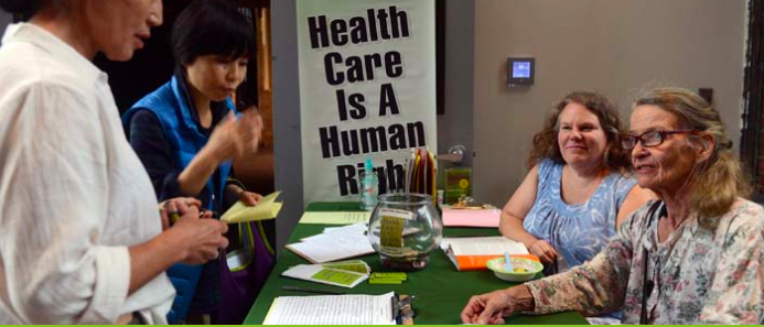 Two women seated at a table during a health event, with informational materials and a clear sign reading 'Health Care Is A Human Right' in the background.