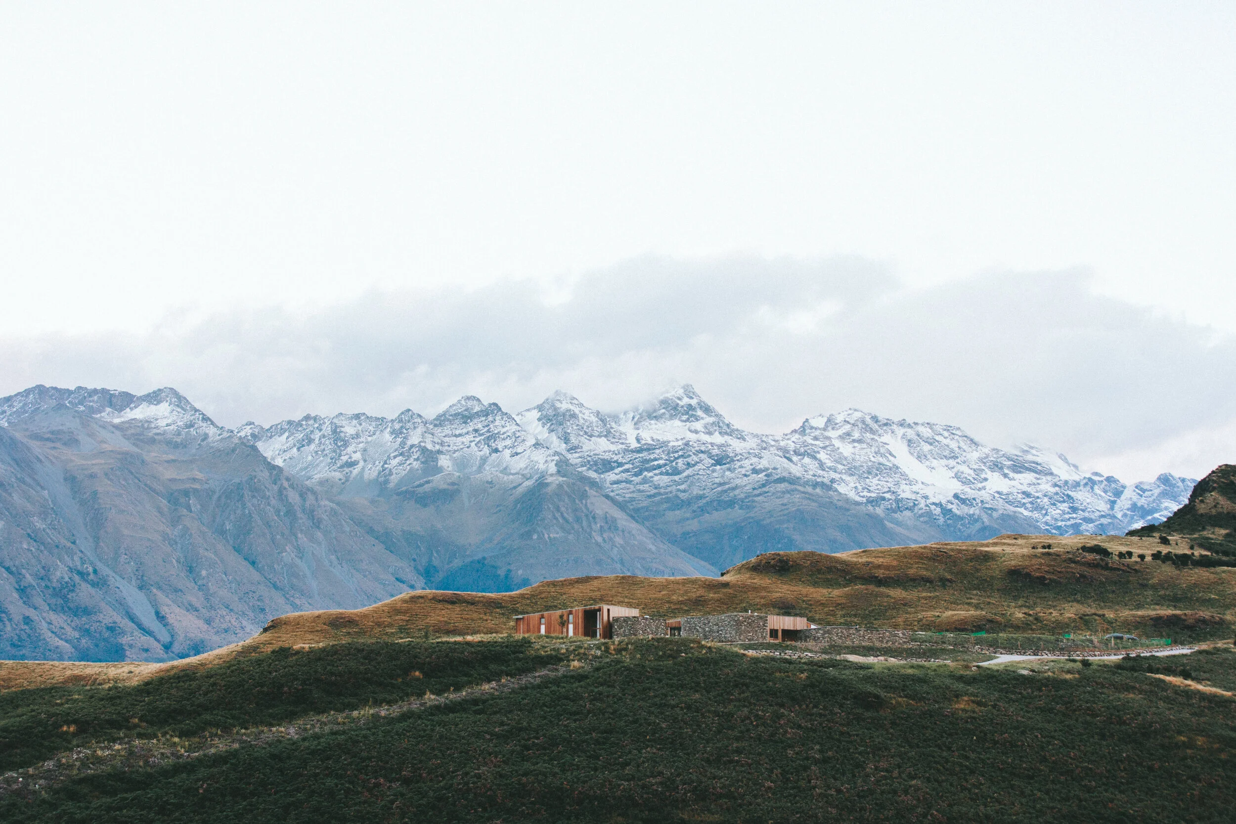 Hügel mit modernen Gebäuden, im Hintergrund schneebedeckte Berge und Wolken am Himmel.