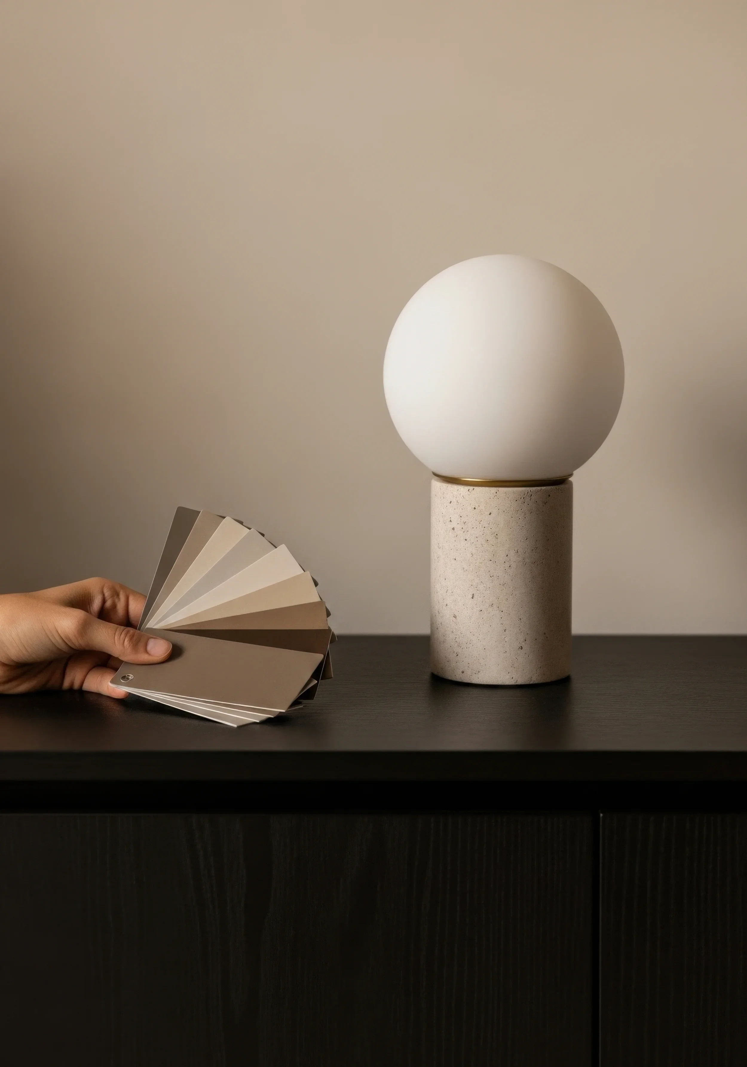 Eine Hand hält eine Fächerkarte neben eine moderne Tischlampe mit Keramikfuß und kugelförmigem Schirm auf einem dunklen Tisch.