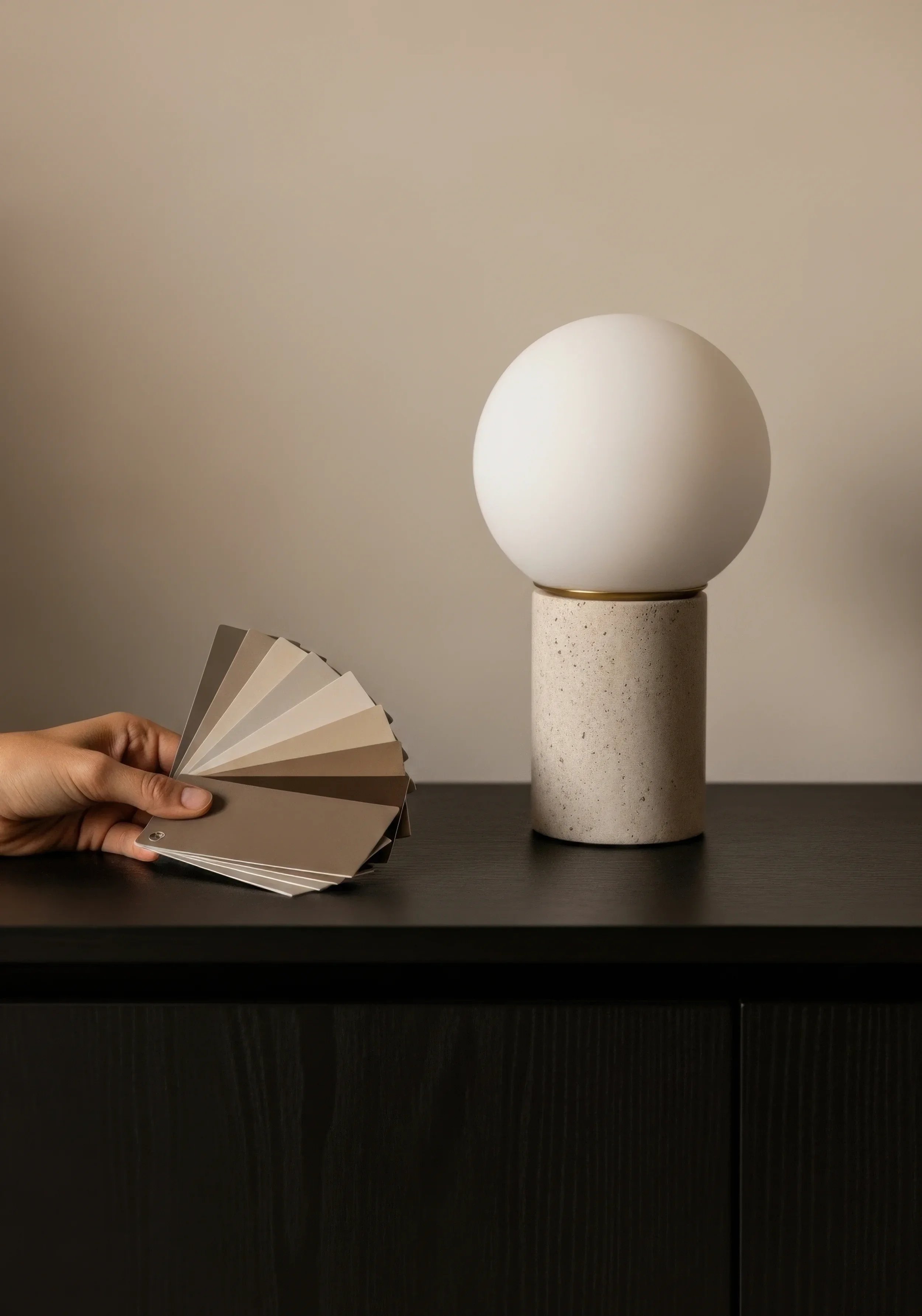 Muestra una mano que sostiene una muestra de varias tonalidades de beige, junto a una lámpara moderna con base de concreto y una esfera blanca, en una superficie negra.
