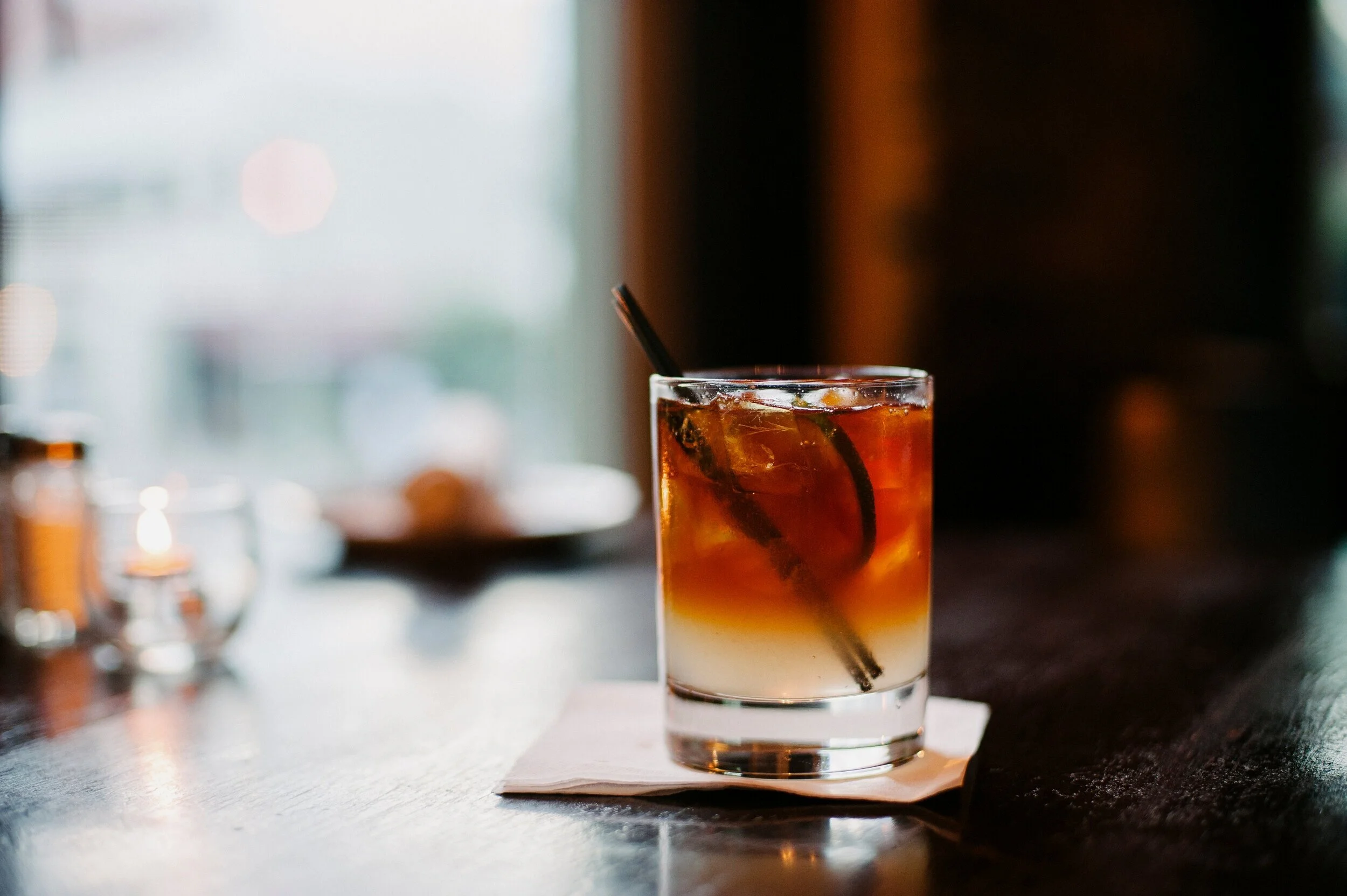 Un bicchiere di cocktail con colore sfumato dall'avorio al rosso, contenente ghiaccio, limone e una cannuccia, posato su un tovagliolo di carta su un banco in un locale