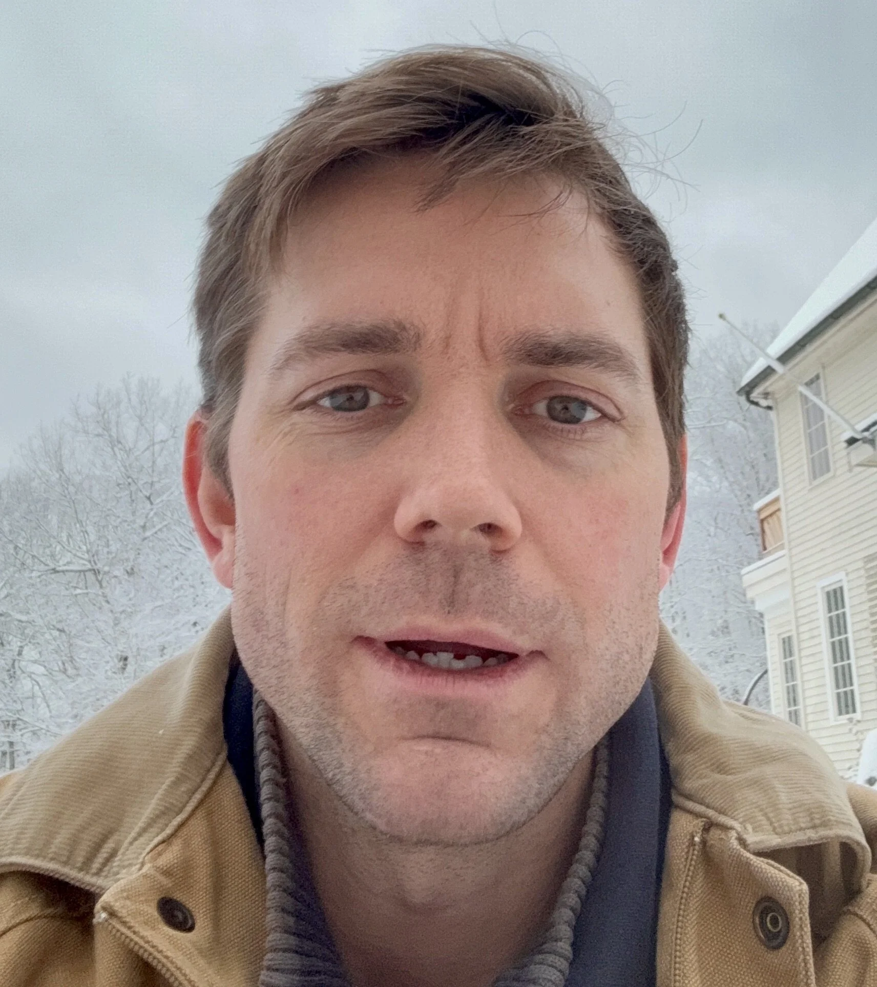 Close-up selfie of a young man with light brown hair, blue eyes, wearing a tan jacket and a hoodie, outdoors with a wintery background