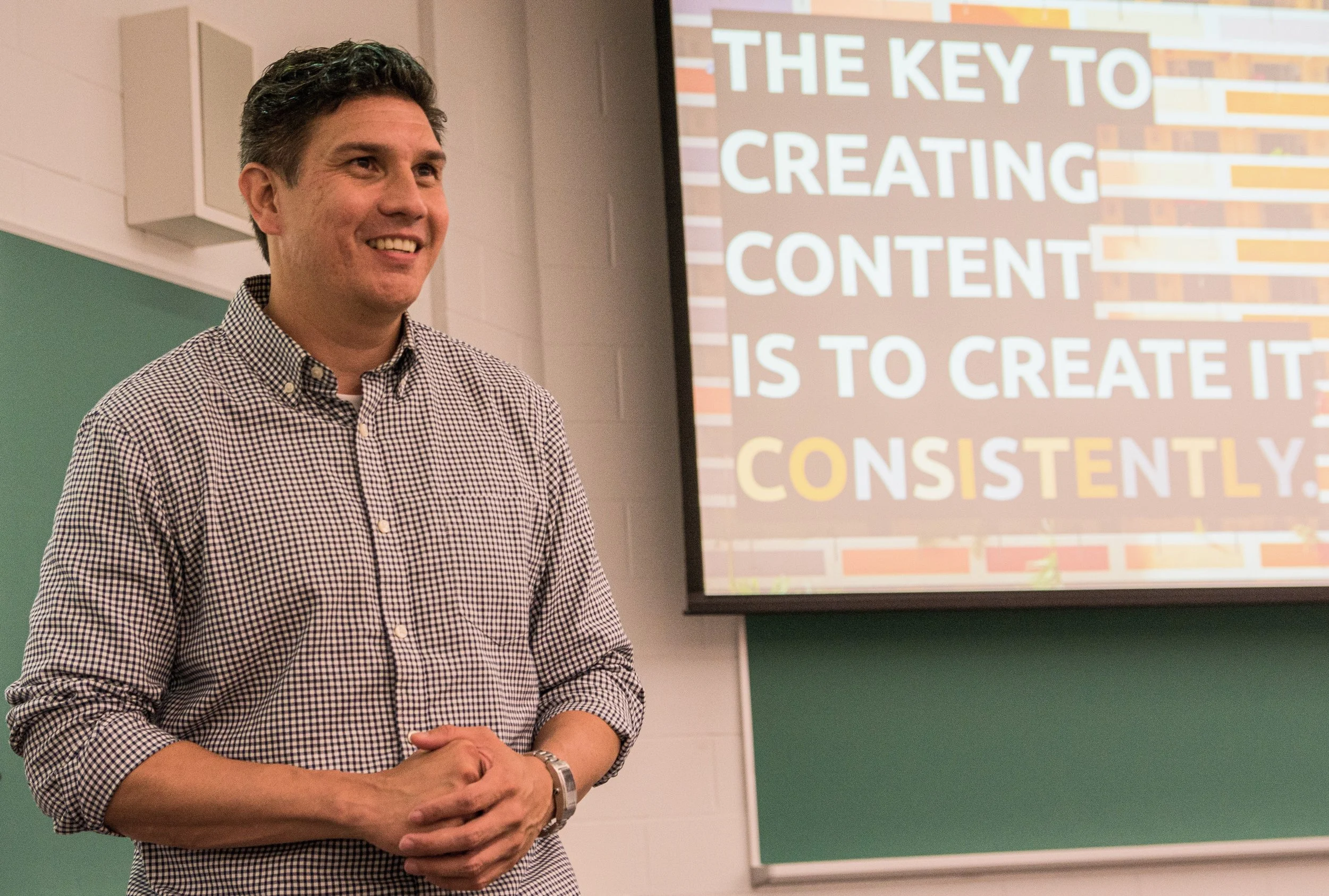 A man smiling in a classroom with a presentation slide about content creation in the background.