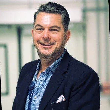 Professional man smiling in an office setting, wearing a dark blazer and blue checked shirt.