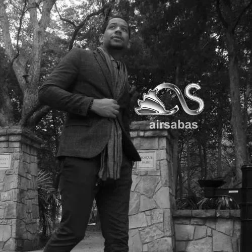 Korey Larkins standing outdoors in a park-like setting with trees in the background, wearing a dark coat and scarf from Suit Supply, with a swan logo and the text 'airsabas' overlayed on the right side.