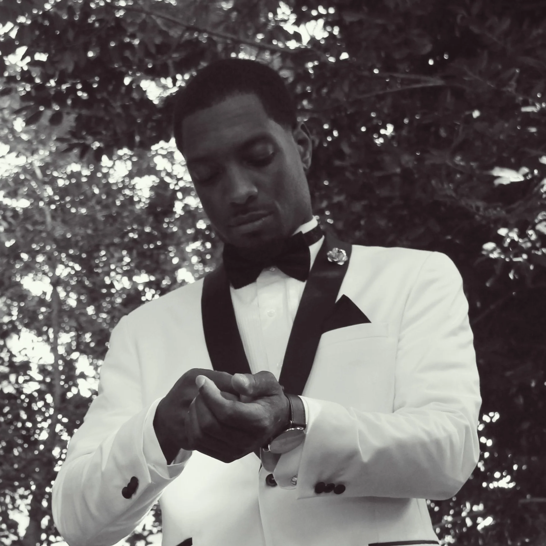 Aubrey Cain dressed in a tuxedo with a white jacket and black lapels, a black bow tie, and a watch on his left wrist, looking down at his watch. The background shows trees with leaves. Spring at The Dallas Arboretum