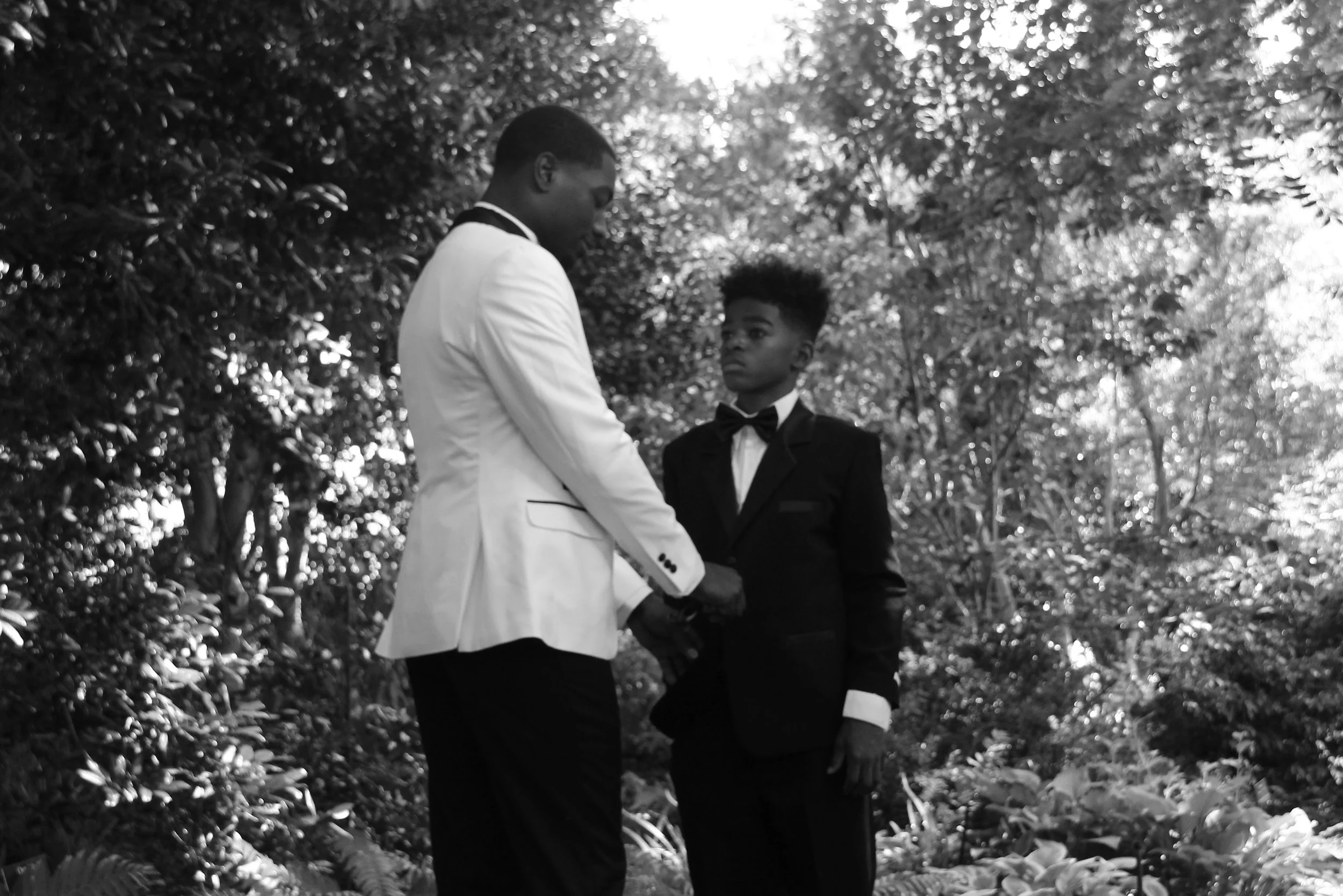Two young men in tuxedos, one in a white tuxedo jacket and the other in a black tuxedo jacket, standing outdoors in a wooded area, holding hands and facing each other.