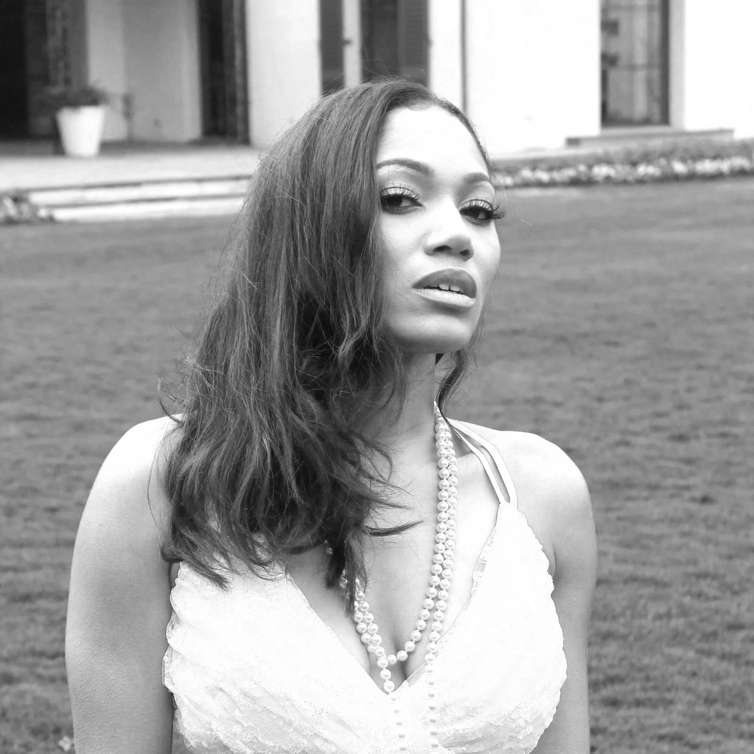 Ty Cain with long wavy hair wearing a lace dress and pearl necklace, standing outdoors on grass, with a building in the background.