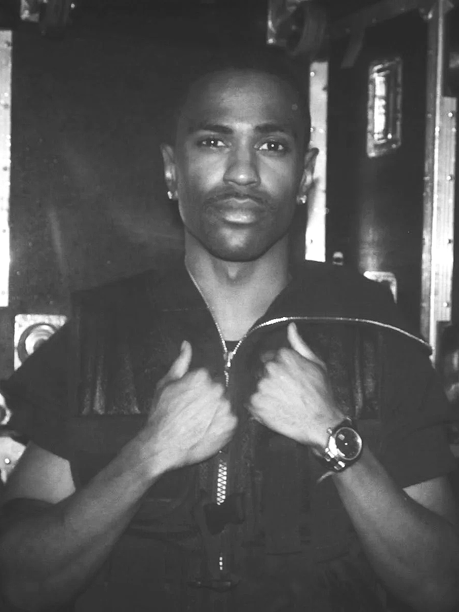 Big Sean with short hair and earrings, wearing a watch, holding the collar of his tactical jacket, posing backstage at the\MTV  Woody Awards.