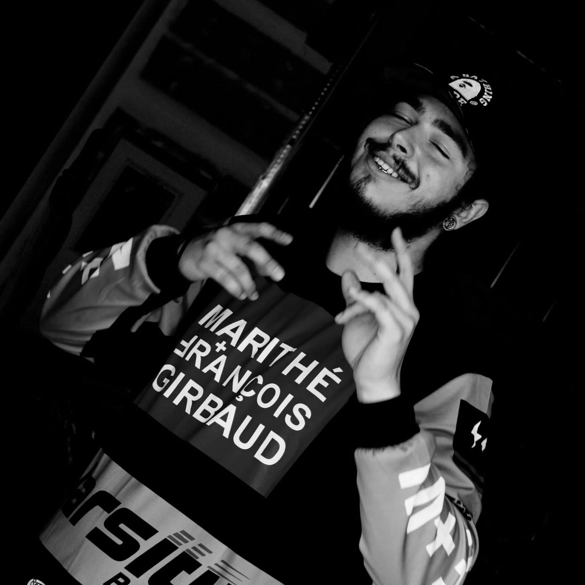 Post Malone with a beard and earrings wearing a cap and a Marithe + François Girbaud jacket, smiling and singing, at a Studio in Dallas.