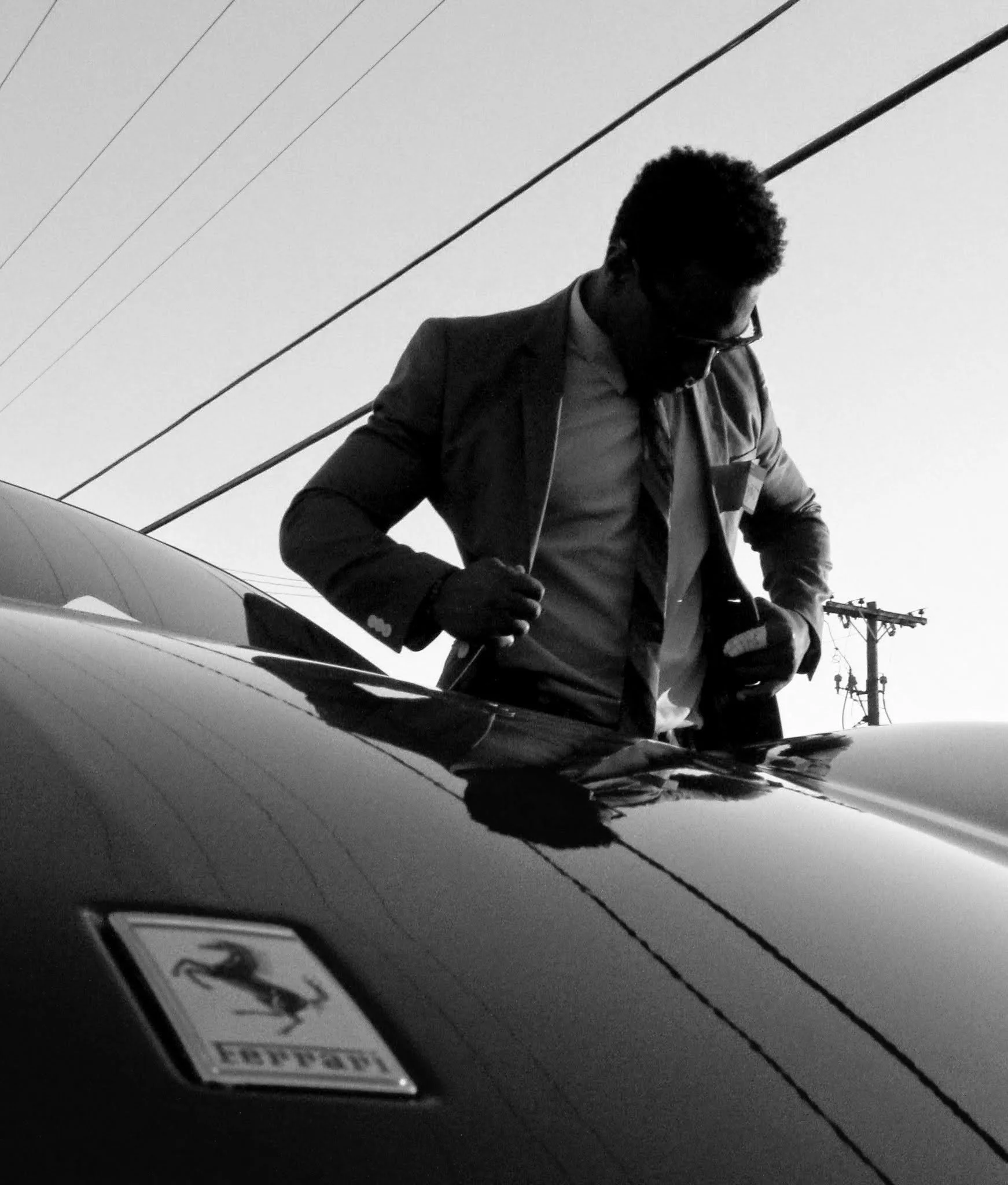 A man in a suit and tie adjusting his jacket near a sleek Ferrari car with the logo visible on the hood, outdoors with power lines in the background.