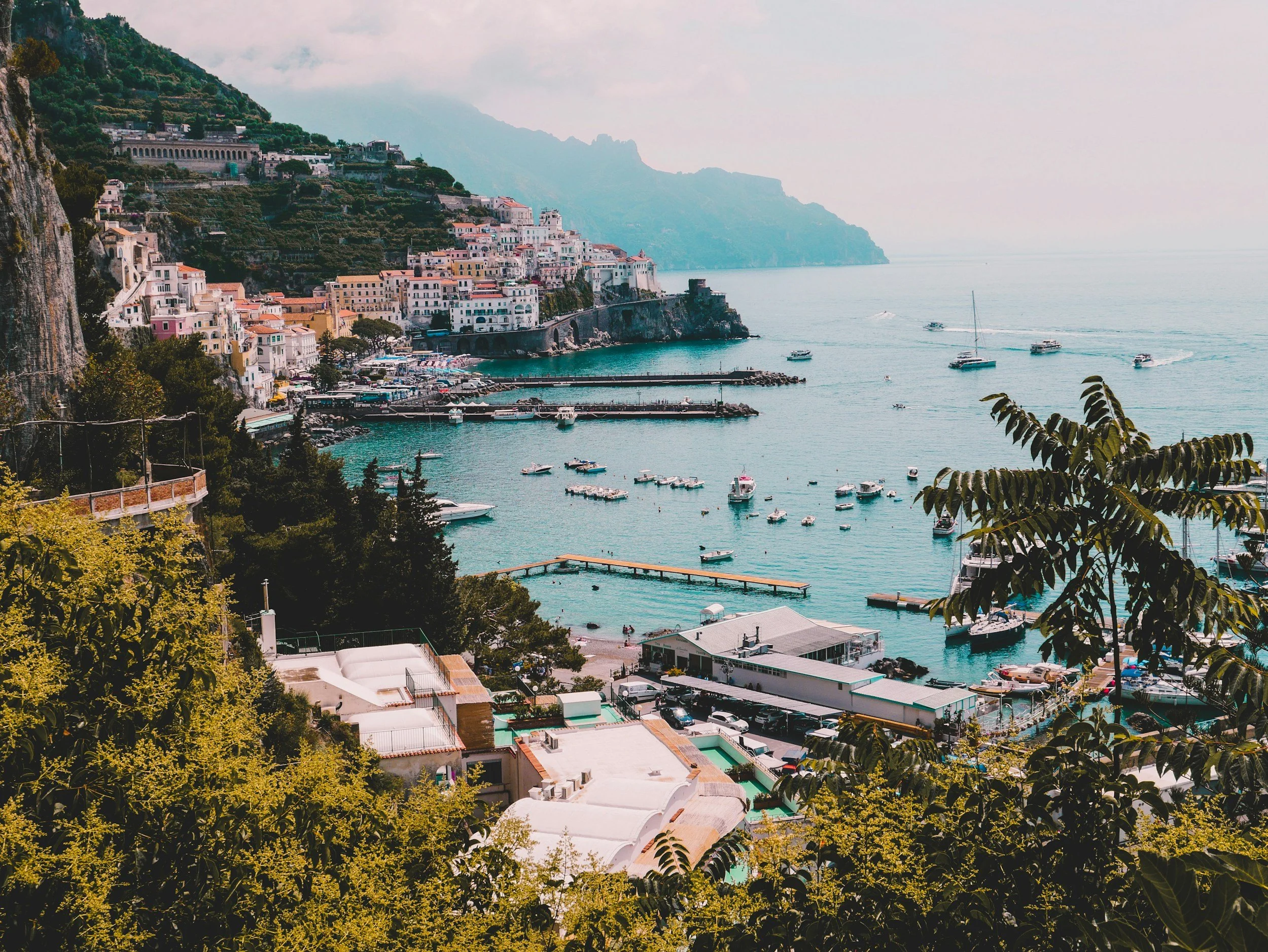 Aerial view of Amalfi's cliffside town and harbor on Italy's Amalfi Coast, with colorful buildings, mountains, and boats in turquoise water