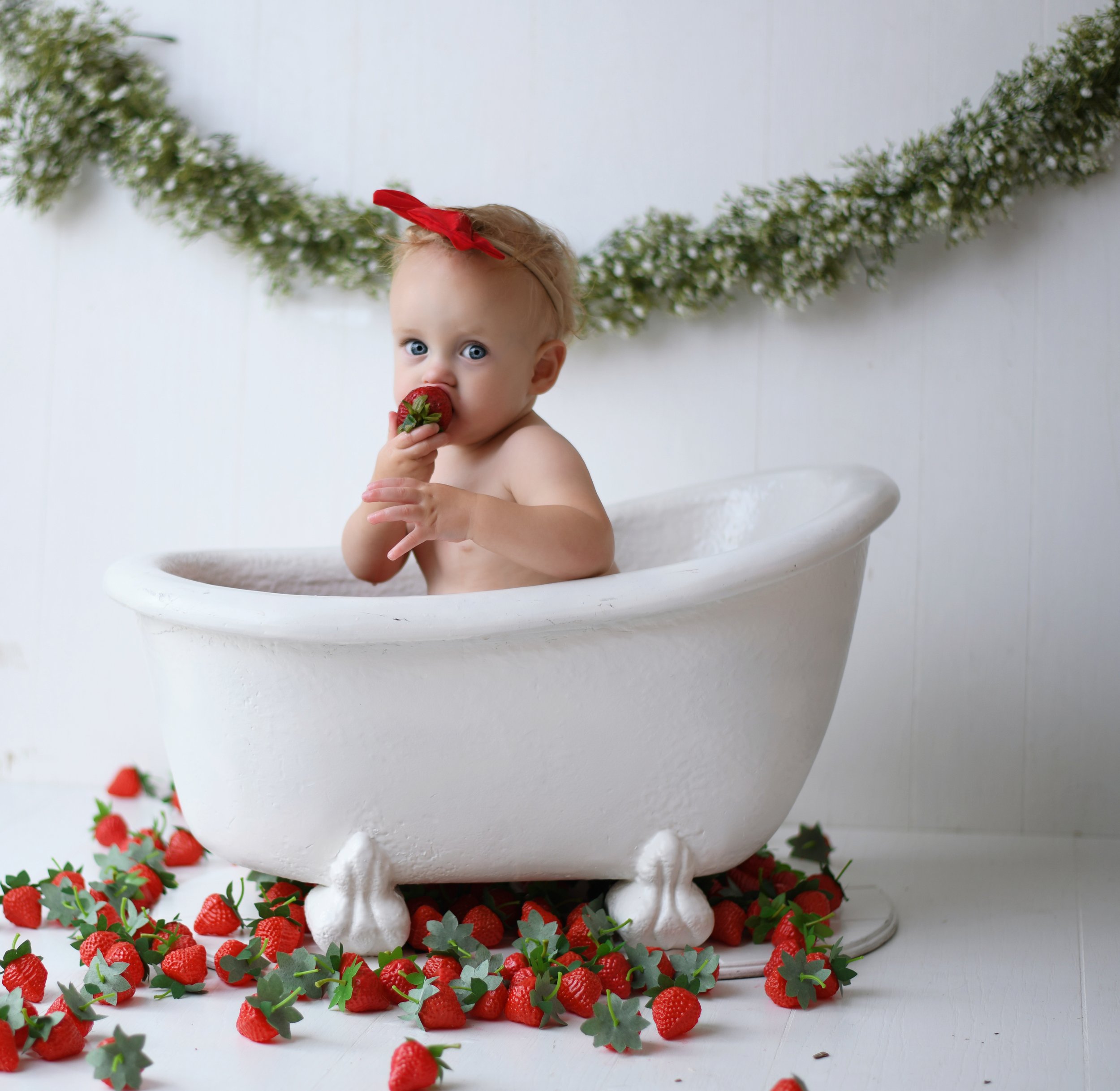 A young child with blonde hair, blue eyes, and a red bow in her hair sitting in a white vintage clawfoot bathtub, eating a strawberry. The scene is decorated with strawberries and greenery on the floor and a green garland on a white wall behind her.