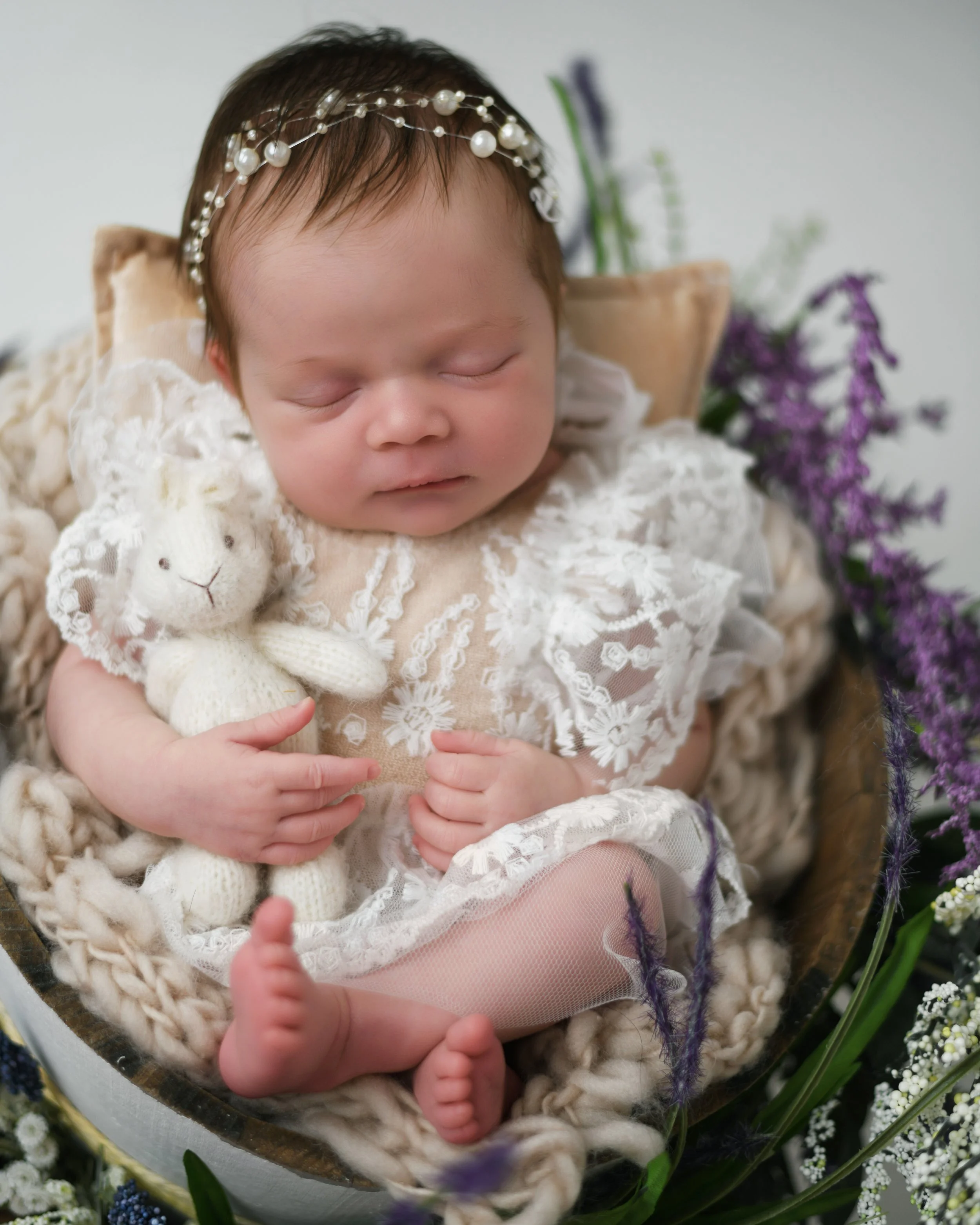 A sleeping baby dressed in an embroidered white dress, holding a small knitted stuffed lamb, with a pearl headband, surrounded by flowers.