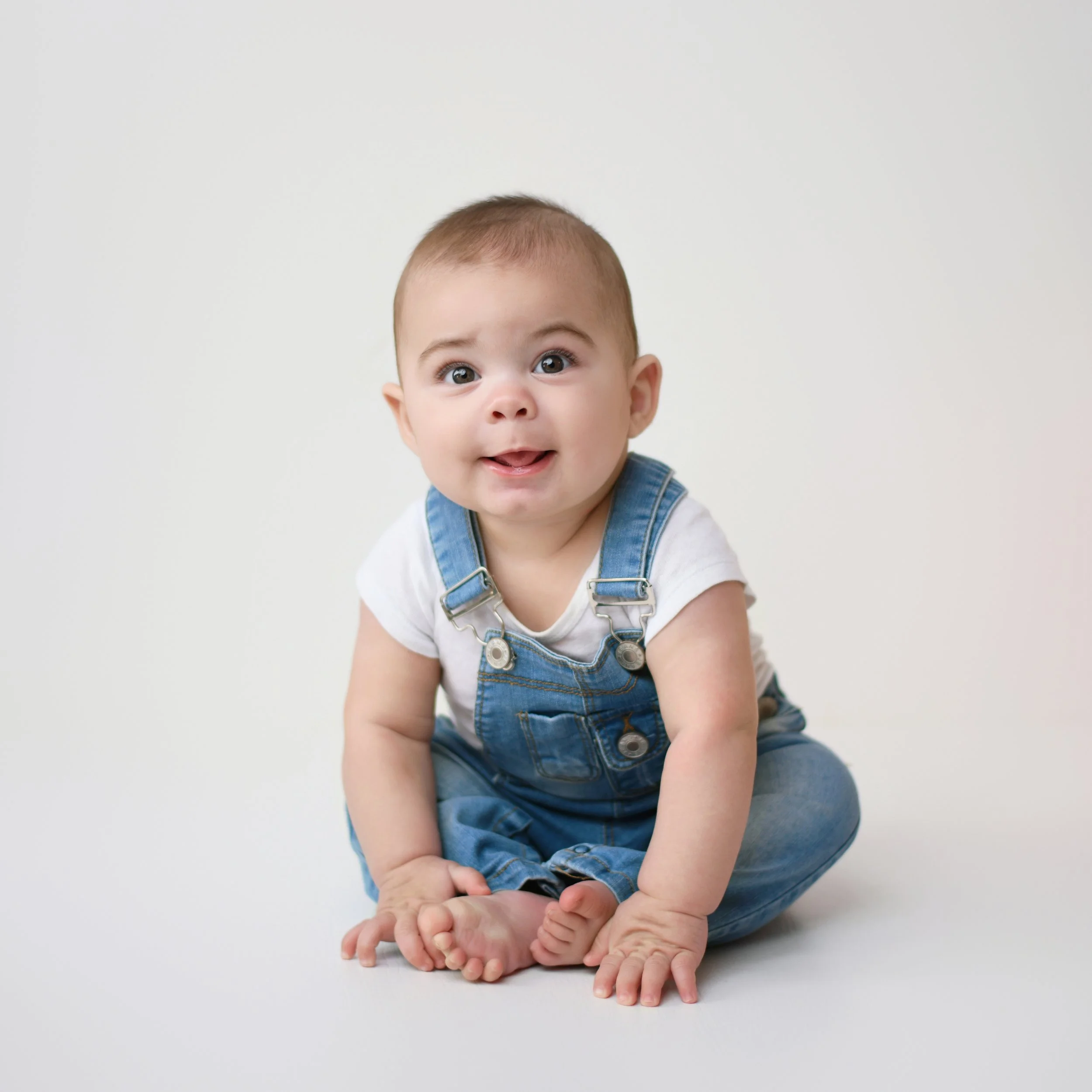 A smiling baby with light skin, wearing blue denim overalls and a white shirt, sitting on the floor against a plain white background.