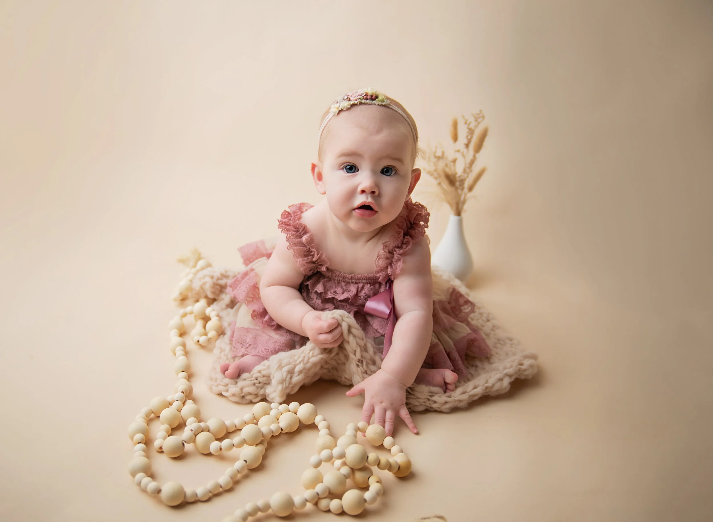 A baby girl in a pink ruffled dress with a pink bow, sitting on a soft blanket, surrounded by beige beads and a white vase with dried pampas grass, against a neutral background.