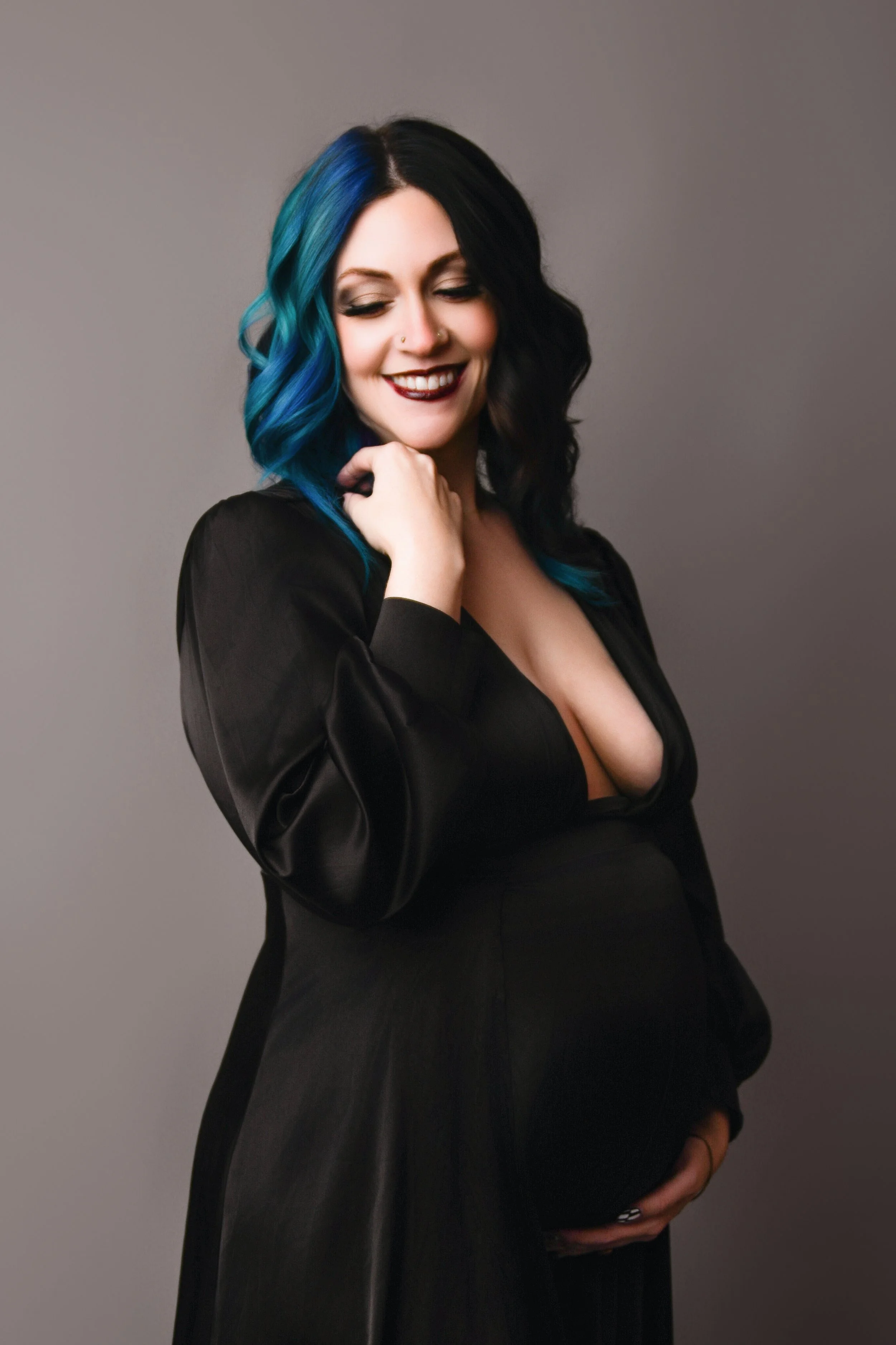 A pregnant woman posing with her eyes closed, smiling, wearing a black dress with a deep neckline, with hair dyed black and blue, against a plain gray background.