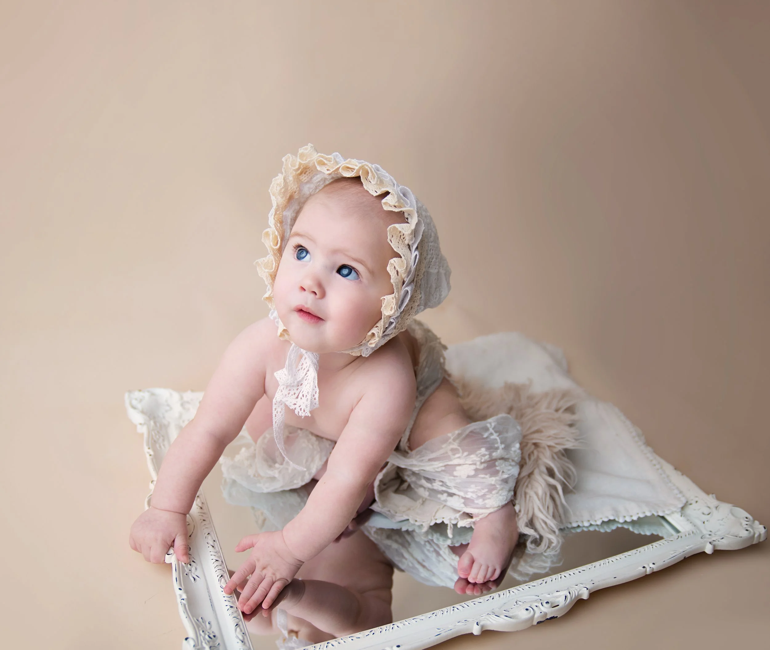 A baby with blue eyes wearing a vintage lace bonnet and matching lace garment, crawling on a decorative mirror with a beige background.