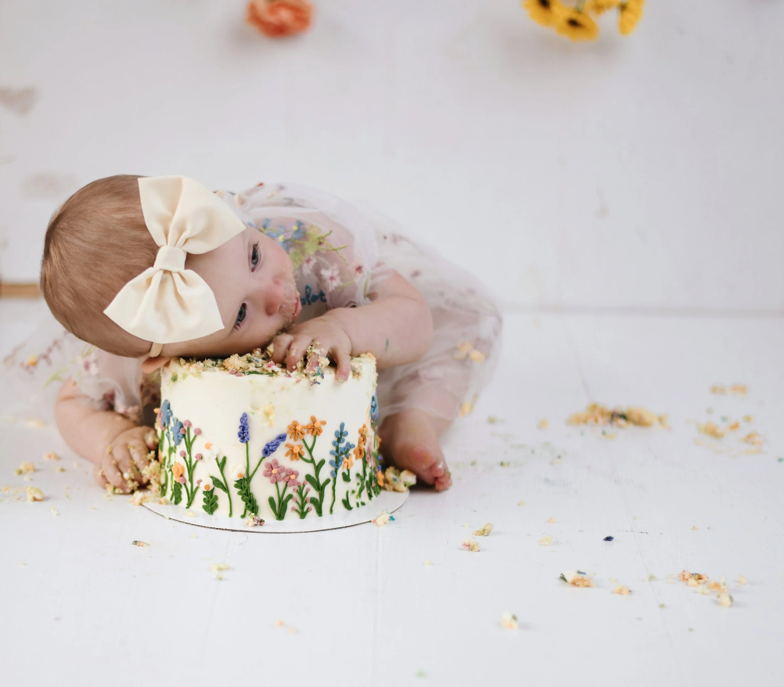 A young girl with a large cream-colored bow headband eating a cake decorated with colorful flowers while sitting on the floor, surrounded by cake crumbs.