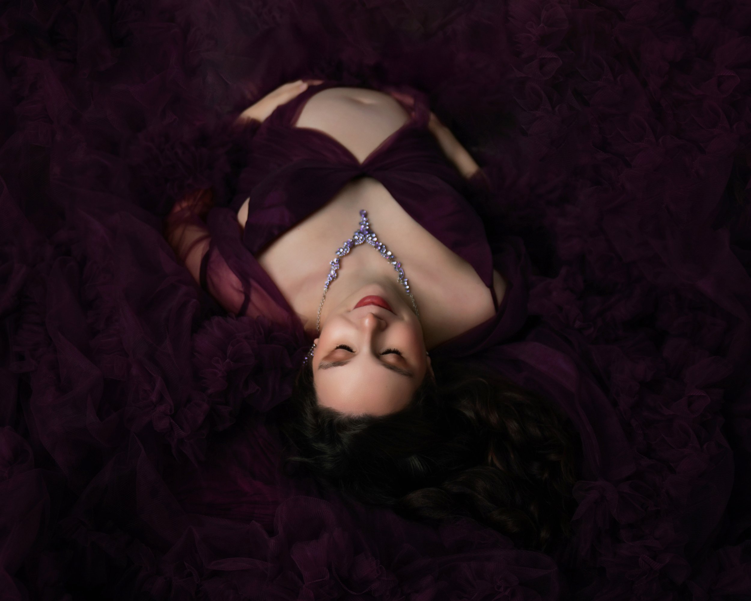 A woman lying on her back wearing a maroon dress with ruffled fabric, a rhinestone necklace, and her eyes closed with her head tilted slightly up.